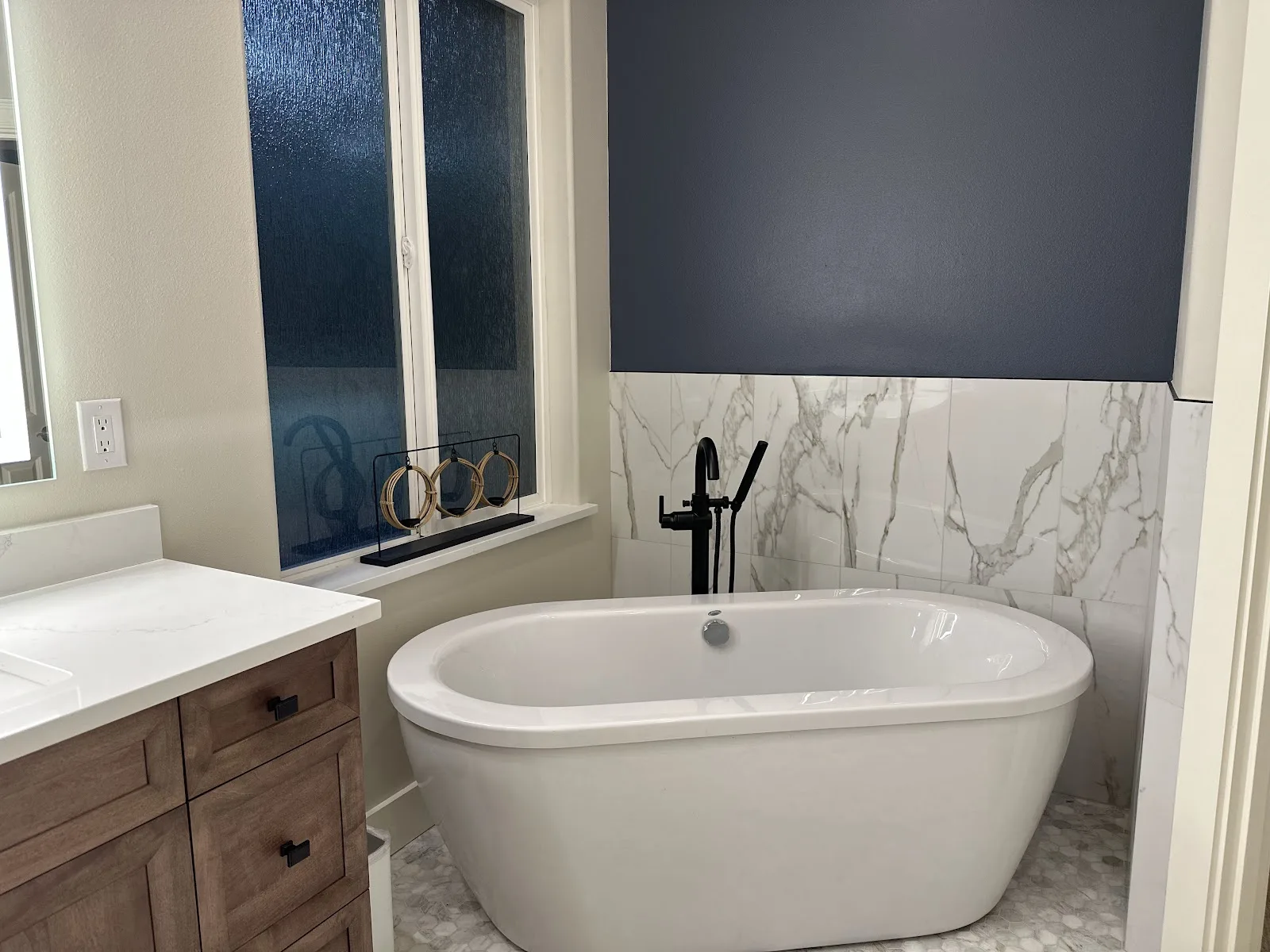 Blaine primary bathroom with navy wall, marble-look tile, wood vanity and freestanding tub in Blaine, WA — Living By Design (3)